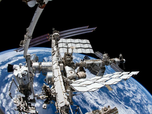 The International Space Station orbits above Earth with blue oceans and white clouds visible below.