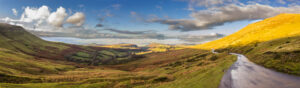 Winding road through sunlit hills and green valleys under a partly cloudy blue sky.