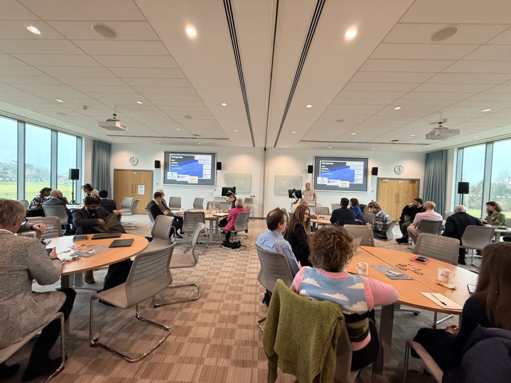 People seated at round tables in a modern conference room, listening to a presentation with slides displayed.