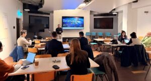 People attend a presentation in a modern meeting room, with laptops open and a speaker at the front.