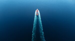 A boat moves through calm blue water, leaving a wide trail of white foam behind it, viewed from above.