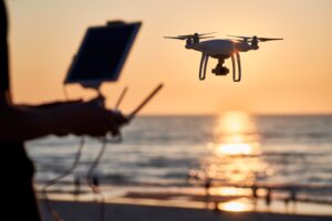 A person controls a drone by the beach at sunset, with the ocean and sun in the background.