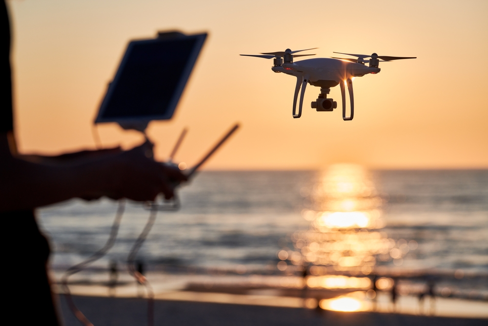 A person controls a drone by the beach at sunset, with the ocean and sun in the background.