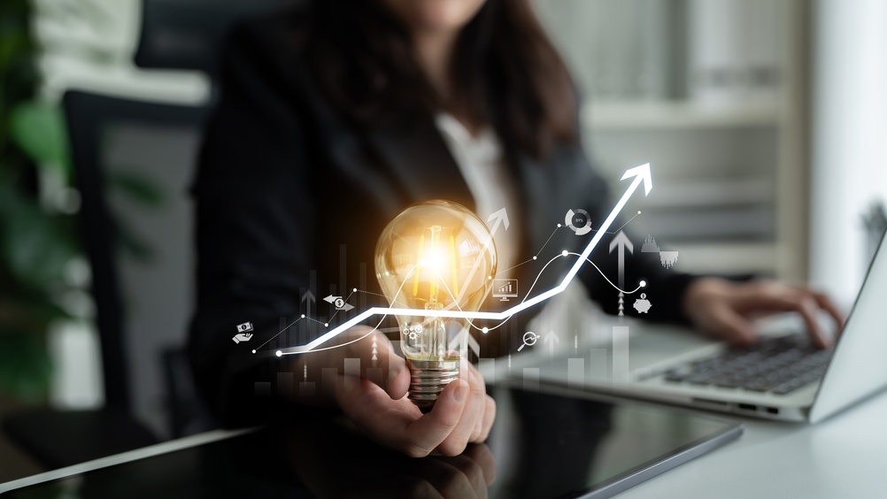 A businesswoman holds a glowing lightbulb with rising graph icons, symbolizing innovation and growth.