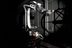 A robotic arm with exposed wires is illuminated against a dark industrial background.