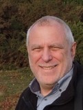 Smiling older man with short gray hair standing outdoors in front of greenery.