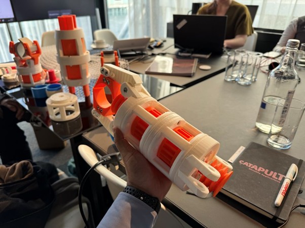 A hand holds a 3D-printed device at a table with laptops, notebooks, bottles, and more 3D models.