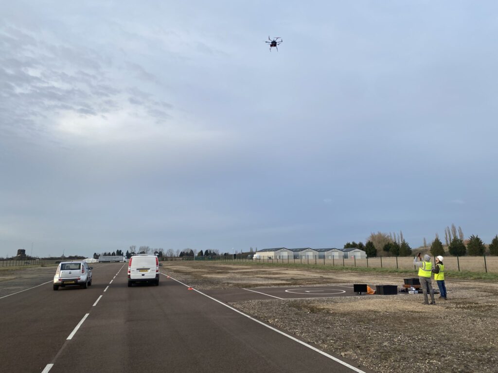 Two people operate a drone near an airstrip, as two vehicles drive on an adjacent road under a cloudy sky.