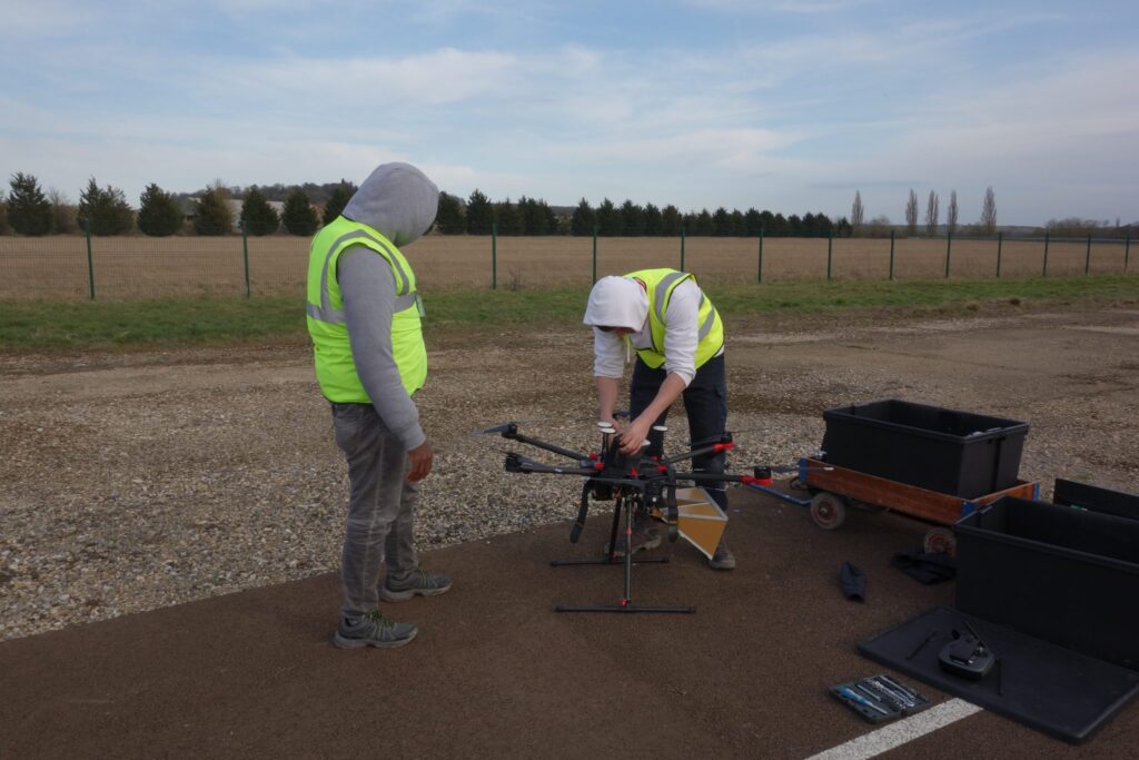 Two people in safety vests prepare a large drone on a paved surface beside black equipment cases.