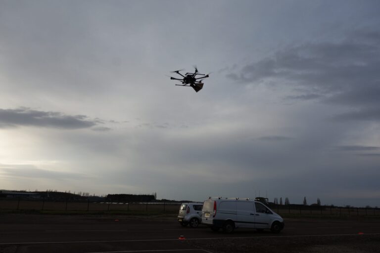 A drone carries a package while flying over two parked vans on a cloudy day near a field.