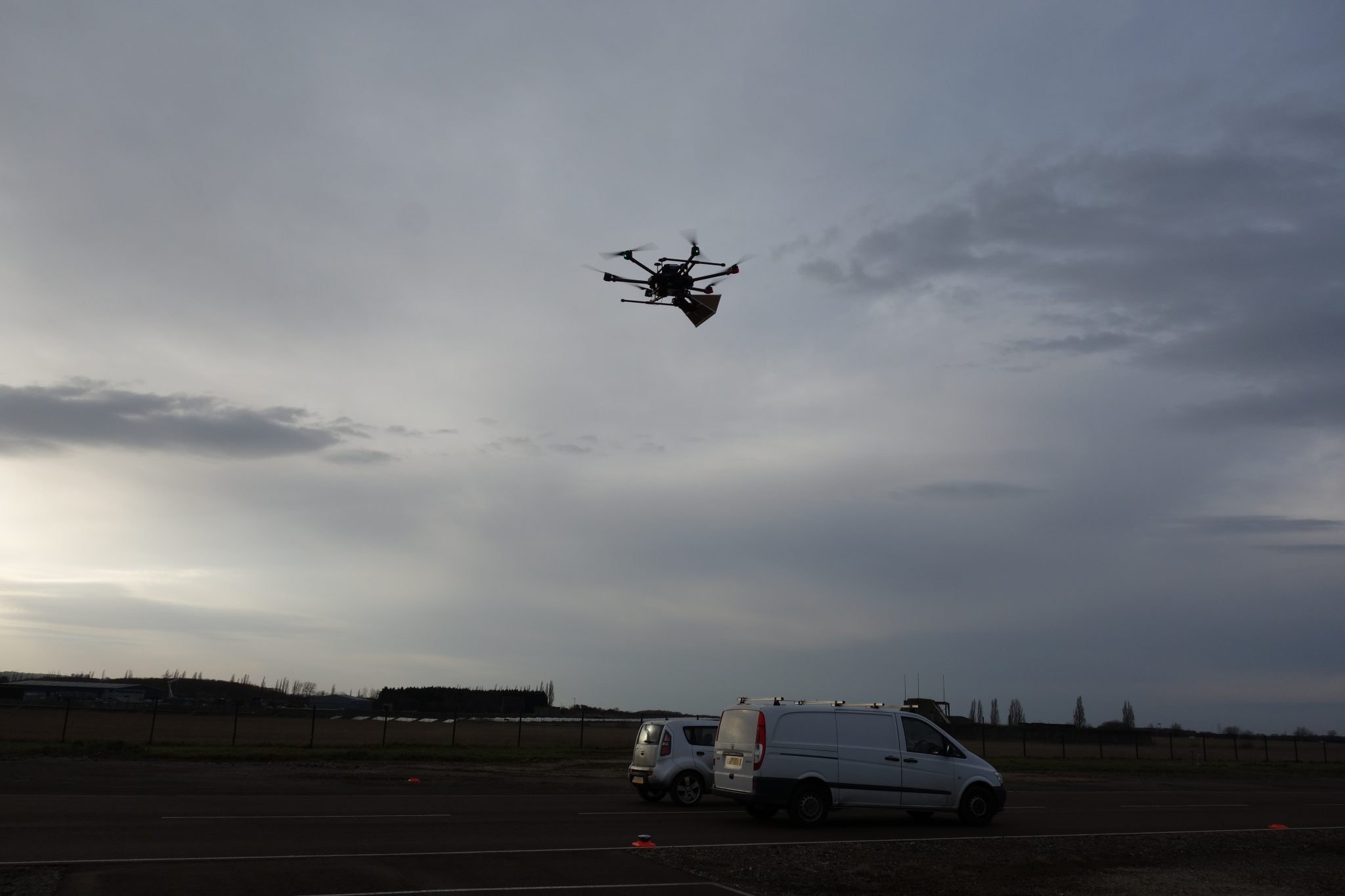 A drone carries a package while flying over two parked vans on a cloudy day near a field.