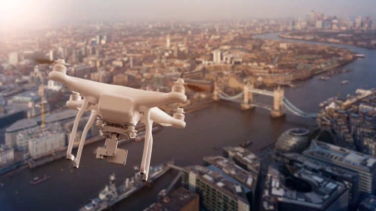 A white drone flying over London with the Tower Bridge and River Thames visible below.