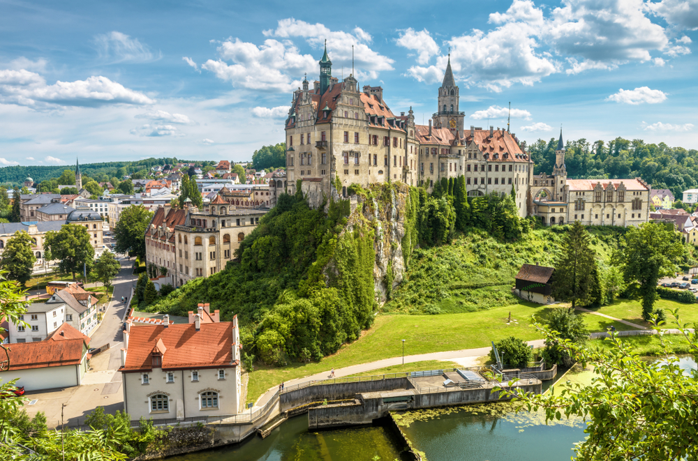 A large historic castle sits atop a green hill overlooking a river and a small town on a sunny day.