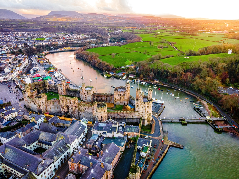 Aerial view of a large medieval castle beside a river, surrounded by a town and green fields at sunset.