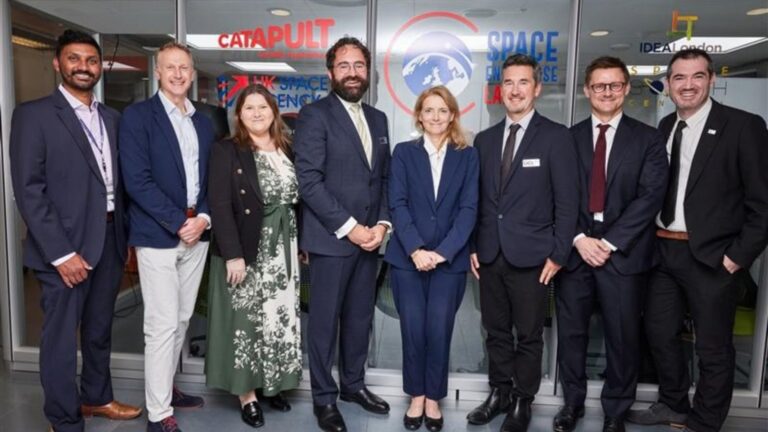 Eight people in business attire stand inside an office in front of a wall with business logos.