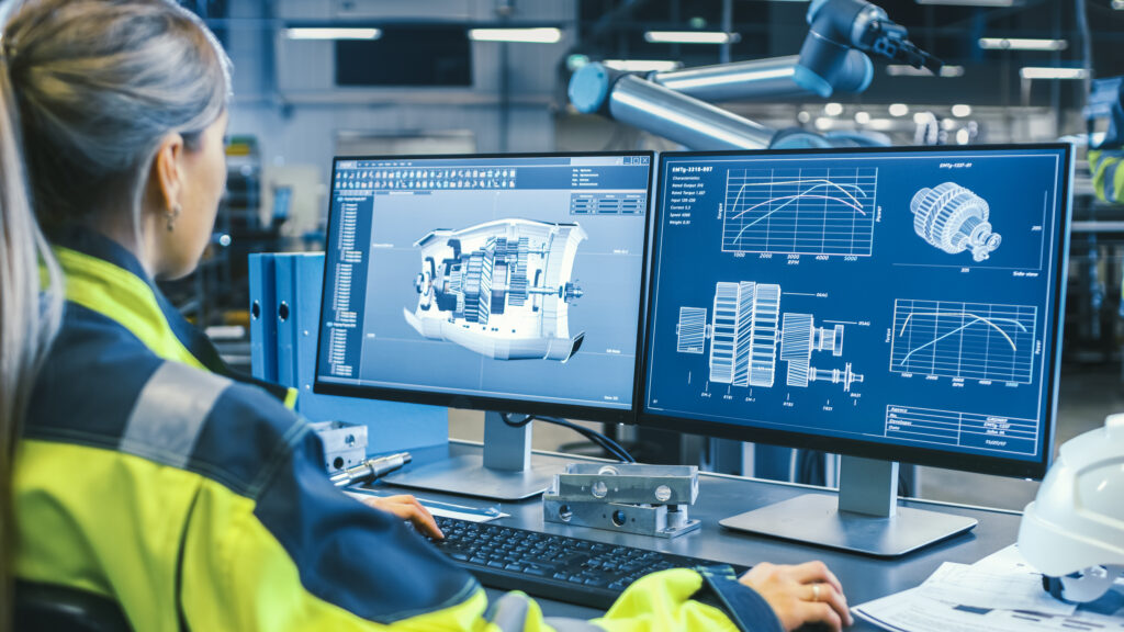 Engineer reviews 3D mechanical component models and technical graphs on dual monitors in an industrial office.