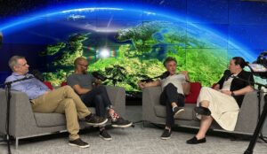 Four people seated in armchairs for a podcast recording in front of a large screen showing an image of Earth from space.
