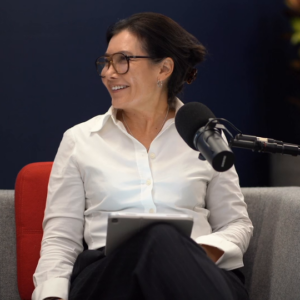 A woman wearing glasses and a white button-down shirt sits on a sofa, speaking into a microphone.