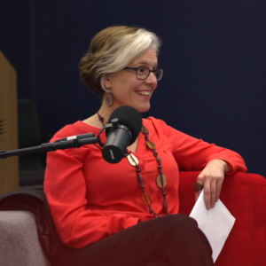 A woman in a red shirt sits in a red chair and speaks into a microphone.