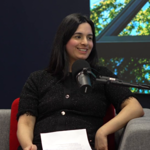 A woman sits in a studio chair, speaking into a microphone while holding a paper.